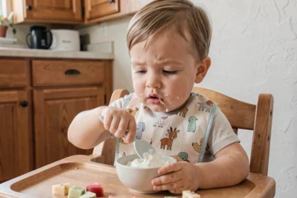 Tout-petit d'environ 18 mois mangeant seul avec une cuillère en silicone dans une chaise haute en bois, apprentissage de l'autonomie alimentaire